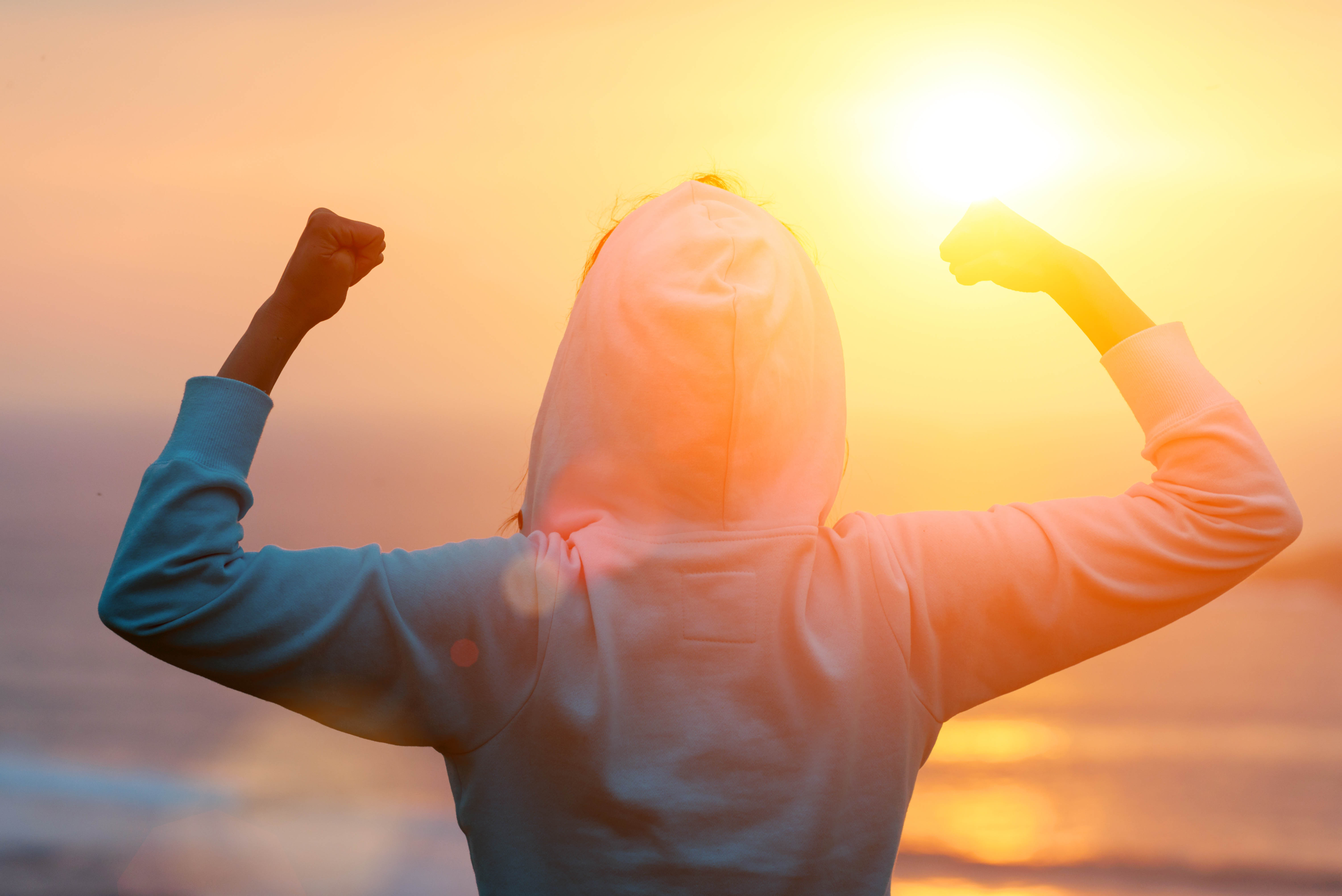 Woman facing the sunset/sunrise, flexing her arms, showing how strong she is